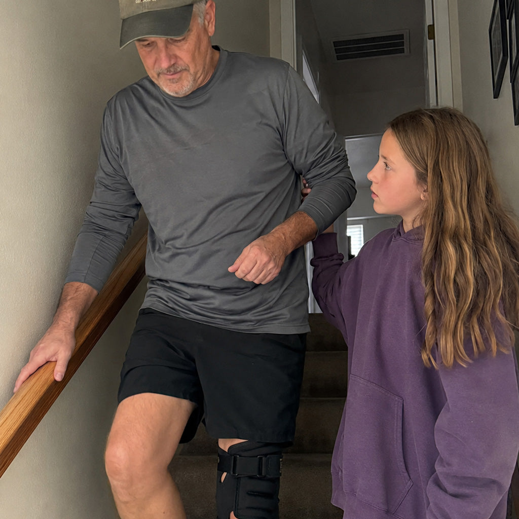 A father makes his way down the stairs gripping the rail while his young daughter steadies his elbow.