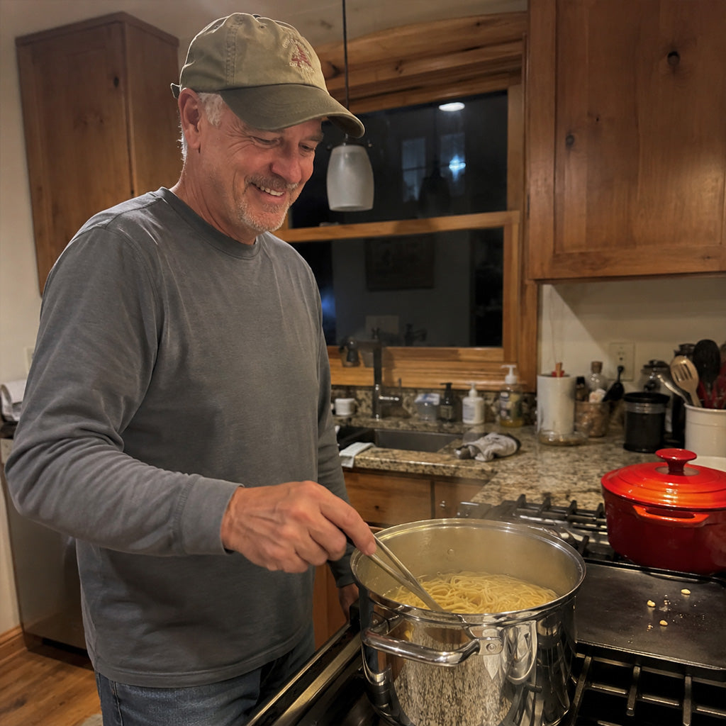 A father stands at the stove cooking spaghetti while his kids do homework at the table behind him.